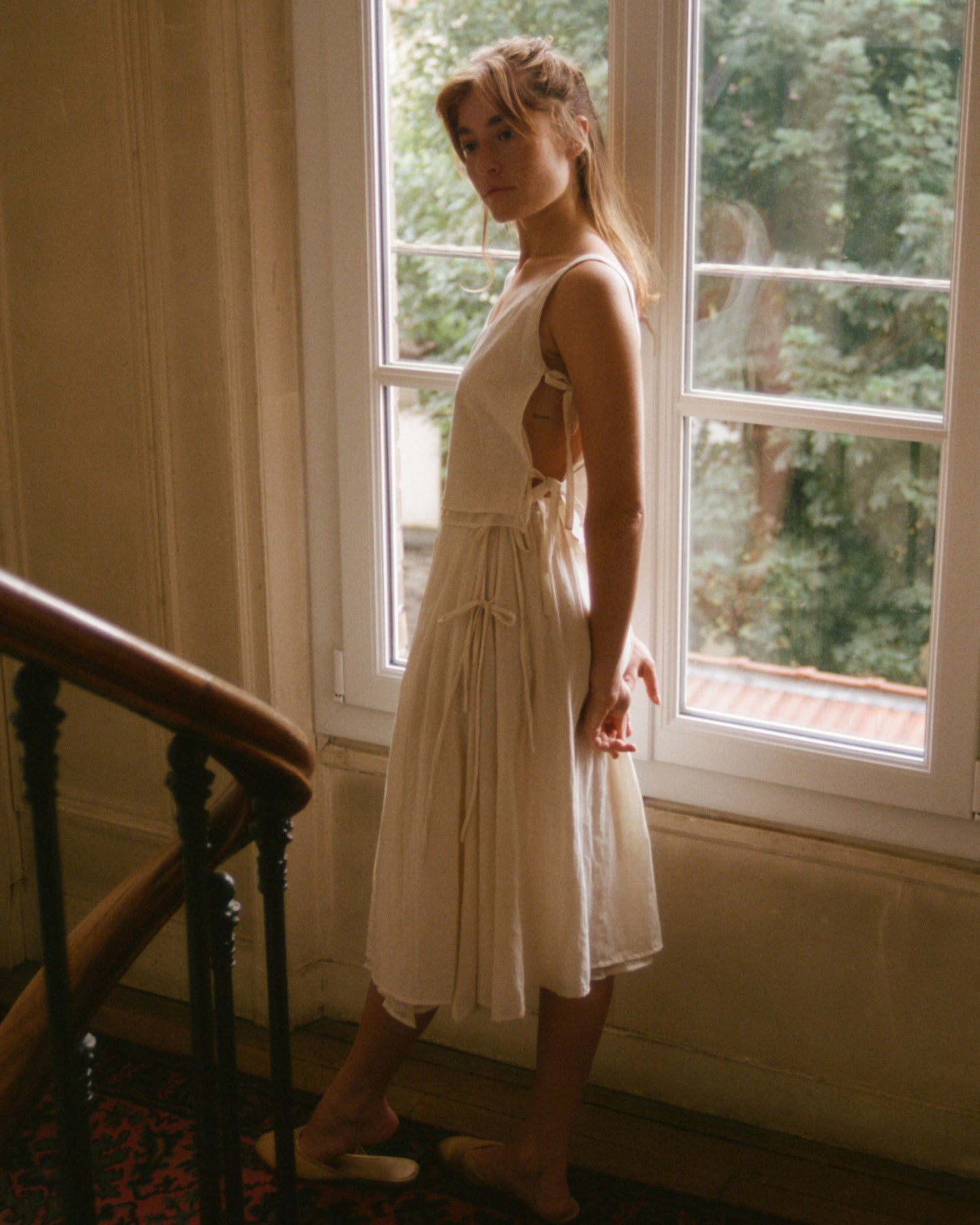 Model by a window wearing Pleated Skirt .01 paired with Lacets Top .01, the perfect summer set, made in Paris.